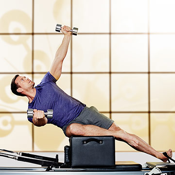 A man in a blue shirt and grey shorts sitting on a Pilates Reformer Box, performing an oblique exercise with weights