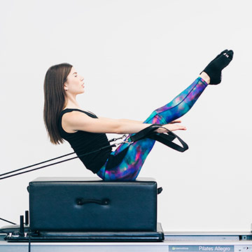 A woman in a black top and blue leggings holding an Pilates Teaser on a Pilates Box